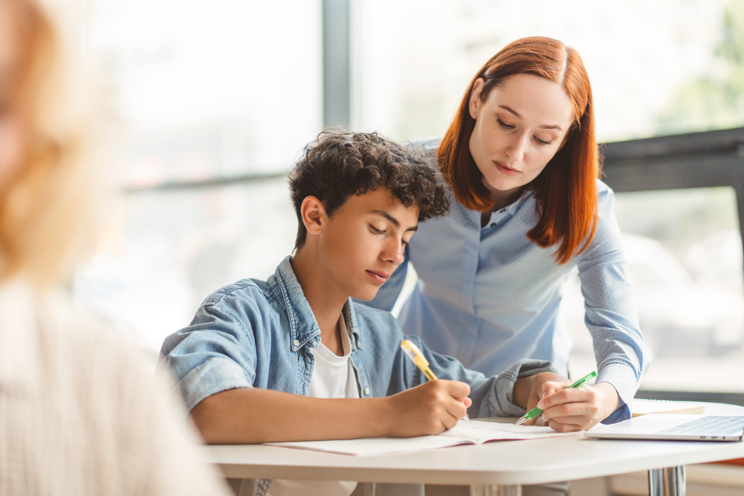 Young pensive school teacher explaining something to smart teenager, standing and showing in notebook, holding pen. Serious tutor assisting helping with homework, exam preparation. Education concept